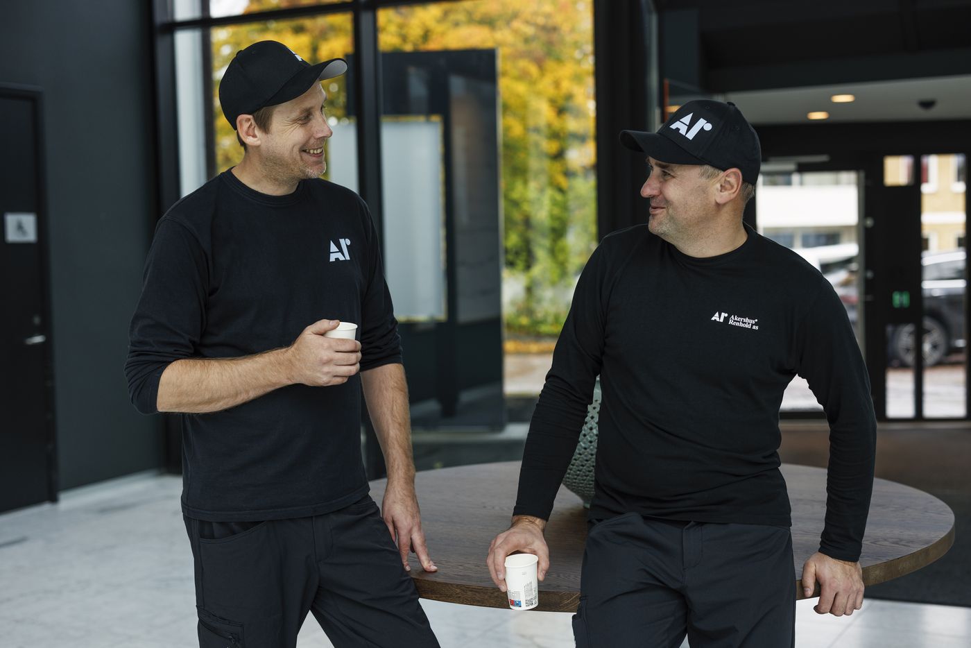 To ansatte i Akershus Renhold-uniform i en lys resepsjon, med kaffe i hånden.